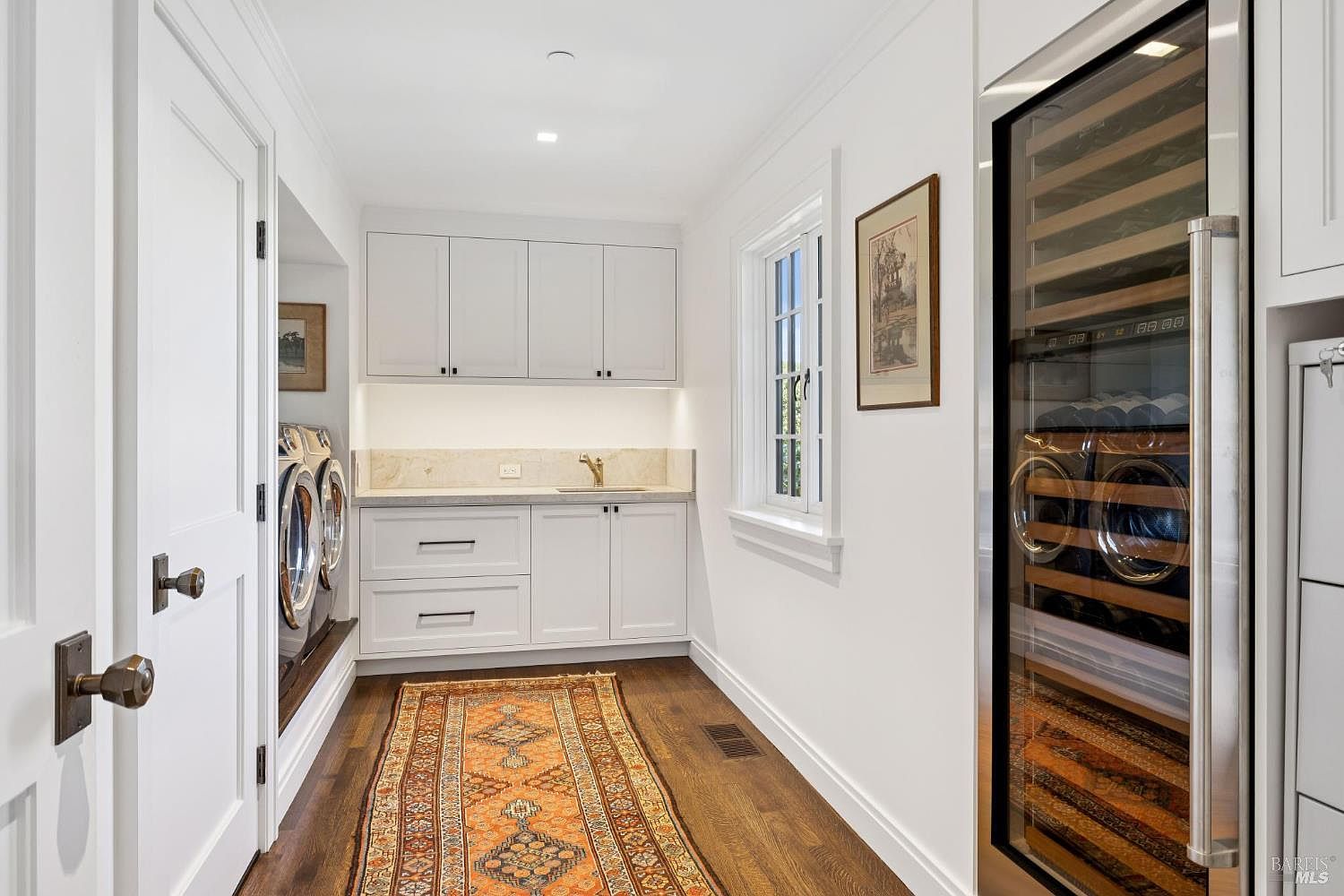 This is a well-organized laundry room featuring white cabinetry, a marble countertop, and a front-loading washer and dryer. An orange patterned rug adds a touch of color to the dark wood flooring. A wine fridge is built into the cabinetry on the right side of the image.