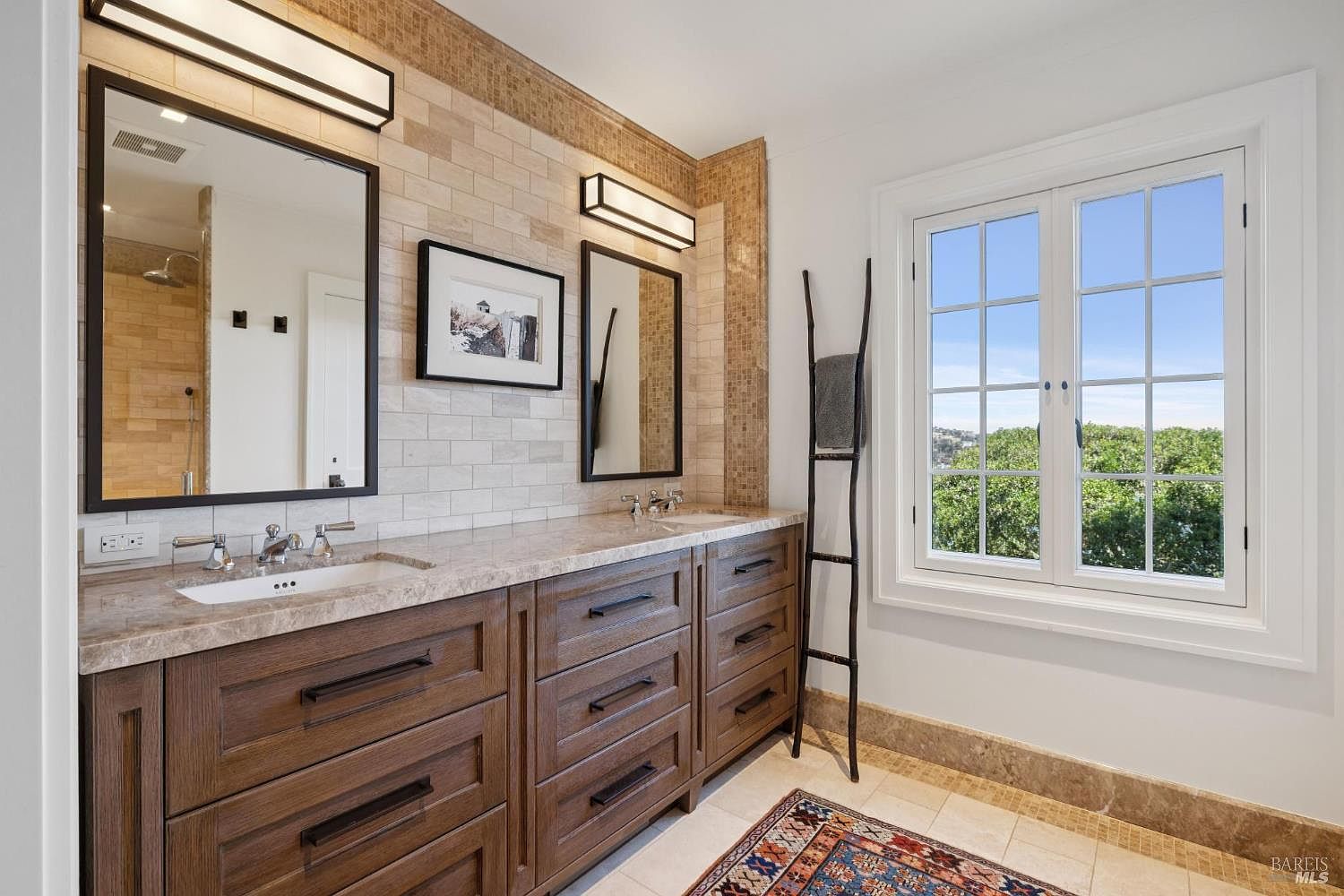 This is a primary bathroom featuring a double vanity with dark wood cabinetry and a light-colored countertop. Two mirrors with black frames hang above the sinks, illuminated by modern light fixtures. A decorative ladder stands near a window offering a view of greenery, and a patterned rug adds a touch of color to the tiled floor.
