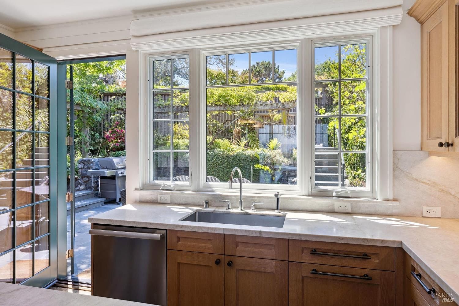 This kitchen features wooden cabinetry with sleek hardware, a stainless steel sink, and a light-colored countertop. A large window above the sink offers a view of a lush garden and outdoor patio area. The open door leads to an outdoor space with a grill, creating a seamless indoor-outdoor living experience.