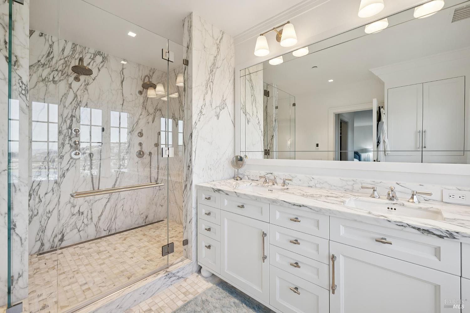 This is a luxurious primary bathroom featuring a glass-enclosed shower with marble walls and a mosaic tile floor. The double vanity has a marble countertop, white cabinetry, and a large mirror with elegant lighting. The overall impression is clean, bright, and upscale, highlighting the high-end finishes and spacious design.