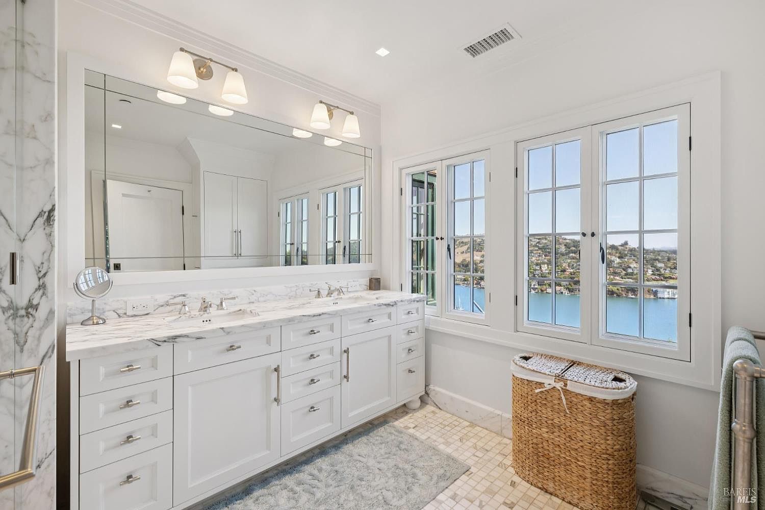 This is a bright and luxurious primary bathroom featuring a double vanity with a marble countertop and white cabinetry. A large mirror spans the length of the vanity, reflecting the natural light from the adjacent windows that offer a scenic view. The room is decorated in a classic style with neutral tones, creating a serene and elegant atmosphere.