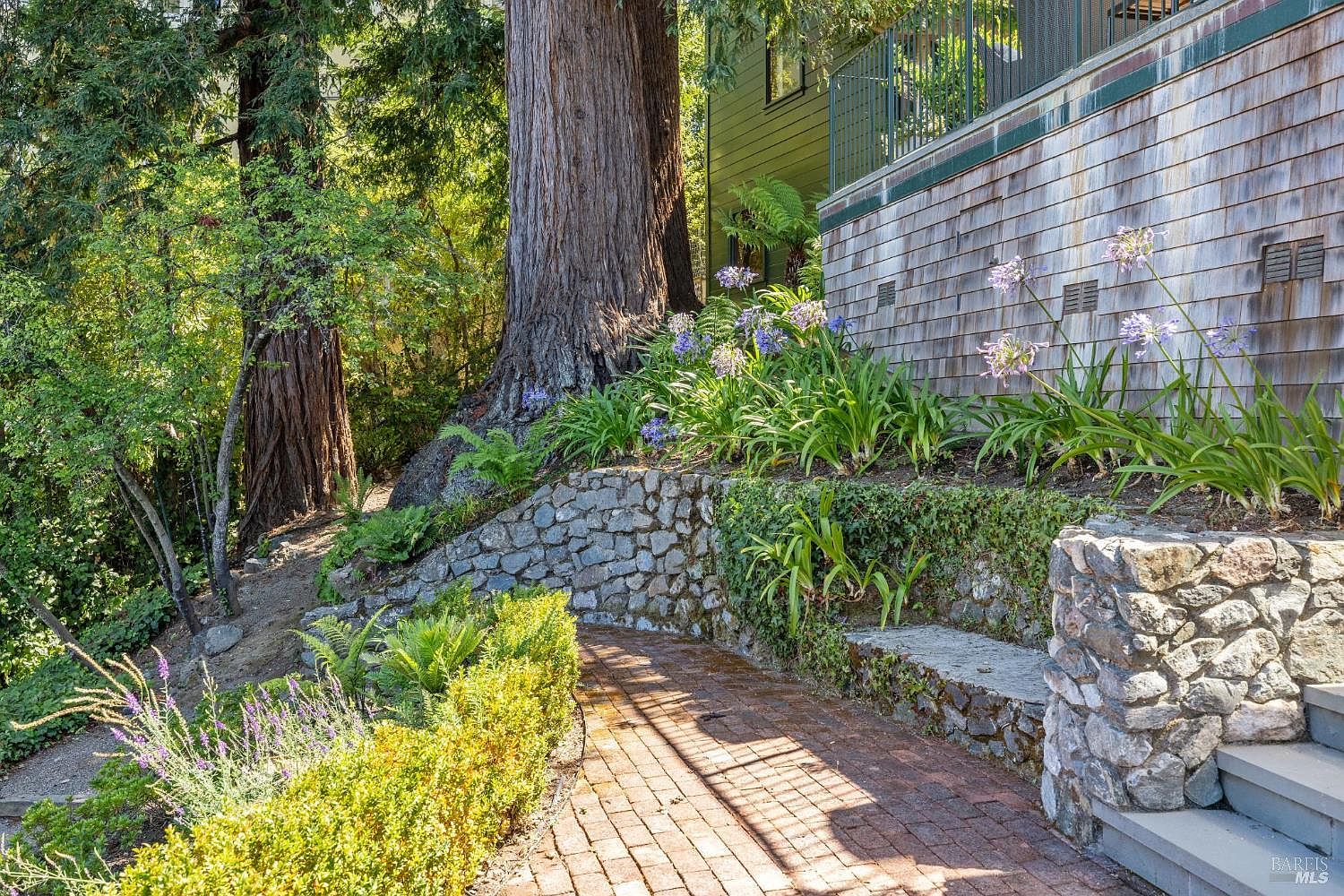 This image showcases a beautifully landscaped yard and garden area. A brick pathway leads through lush greenery, including flowering plants and manicured hedges. A stone retaining wall and mature trees add to the natural charm of the property, creating a serene and inviting outdoor space.