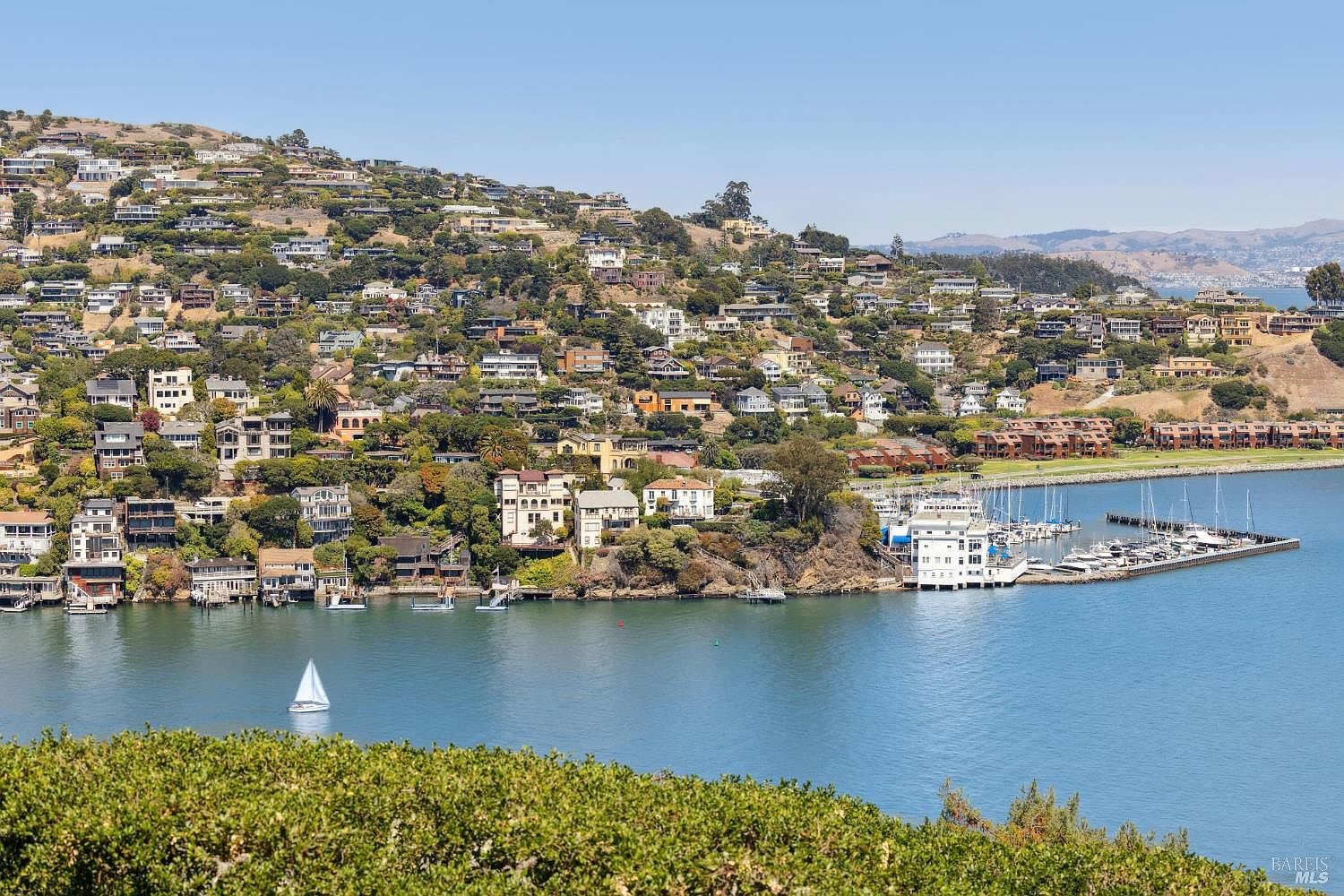 This aerial view showcases a hillside neighborhood with numerous houses nestled among trees, overlooking a bay with a marina. The scene includes a sailboat on the water and a clear blue sky, creating a picturesque and desirable coastal living environment. The marina features several boats docked, adding to the appeal of the location.