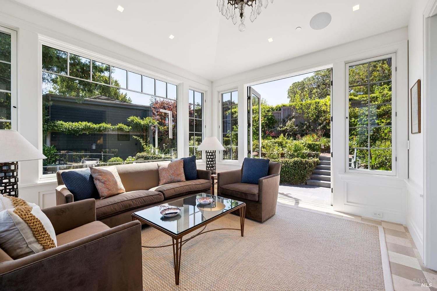This is an interior shot of a sun-drenched living room featuring a neutral color palette. The room is furnished with a brown sofa, two armchairs, and a glass-topped coffee table, all arranged on a light-colored rug. Large windows offer views of the exterior garden, creating a seamless indoor-outdoor living experience.