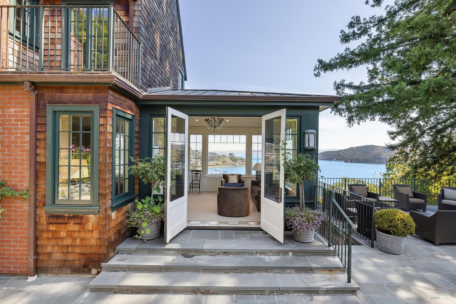 This image showcases a beautiful patio area with a stunning view of the water. The patio features stone flooring, elegant outdoor furniture, and access to an interior space through large, open French doors. The architecture combines brick and wood elements, creating a sophisticated and inviting outdoor living space.
