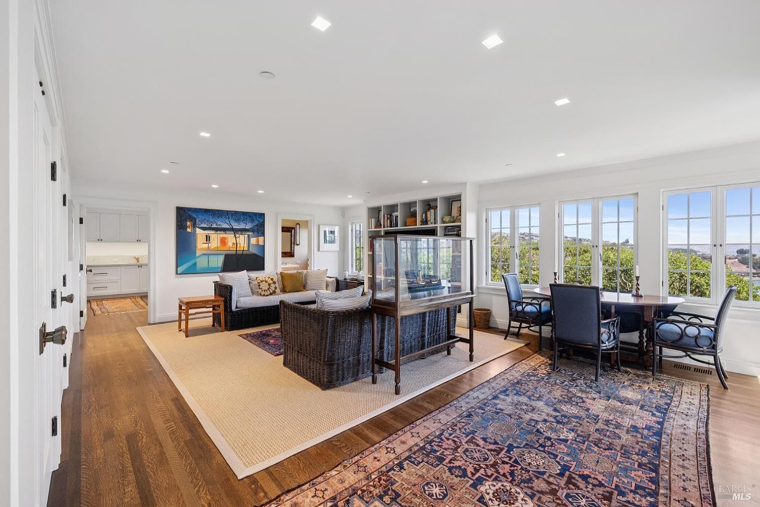 This is an interior shot of a spacious living room featuring hardwood floors, white walls, and ample natural light from large windows. The room is furnished with a sofa, wicker chairs, a display case, and a dining table with chairs, creating a comfortable and inviting atmosphere. An area rug defines the seating area, while a colorful rug adds character to the dining space.