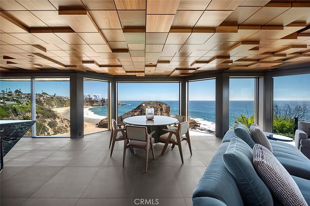 This interior shot showcases a modern dining area with floor-to-ceiling windows offering stunning ocean views. The room features a round dining table with chairs, a unique wooden ceiling design, and a comfortable-looking blue sofa in the foreground. The overall impression is luxurious and serene, emphasizing the property's prime location and high-end finishes.