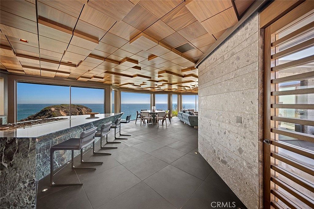 This interior shot showcases a luxurious living space with panoramic ocean views. The room features a unique geometric wood-paneled ceiling, a sleek marble countertop bar with modern seating, and a dining area. A textured stone wall adds depth, while large windows maximize natural light and the stunning coastal scenery.