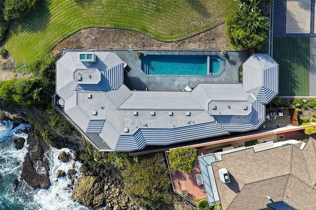 This aerial view showcases a luxurious coastal property with a unique architectural design, featuring a gray roof, a private pool, and meticulously landscaped grounds. The house is perched on a cliff overlooking the ocean, offering stunning views and a sense of exclusivity. The image highlights the property's prime location and upscale amenities.
