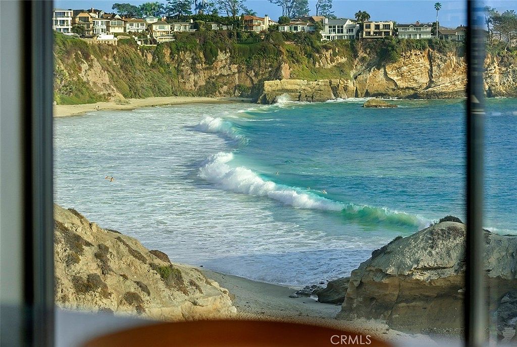 This image showcases a stunning ocean view from what appears to be inside a building, possibly a home or condo. The turquoise water is punctuated by white waves, and a rocky coastline is visible in the foreground and background, topped with houses. The view is framed by a window, suggesting a desirable location with a premium view.