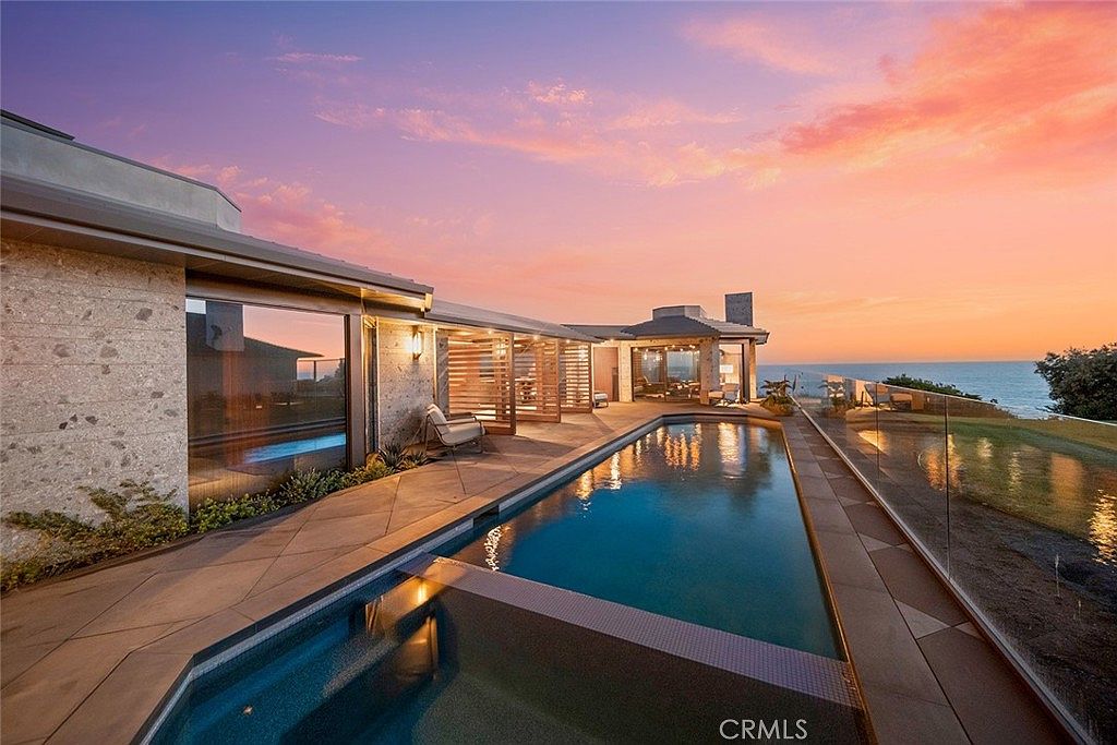 This image showcases a luxurious outdoor pool area at sunset, featuring a sleek, modern design with clean lines and a seamless integration with the surrounding landscape. The pool is surrounded by a stone patio and a glass railing, offering unobstructed views of the ocean. The architecture of the house is contemporary, with large windows and a flat roof, creating a sophisticated and inviting atmosphere.