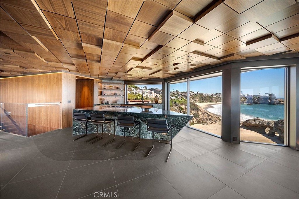 This is a modern kitchen with a unique wooden ceiling design and a large island with a marble countertop and seating. The kitchen features large windows offering a stunning ocean view, enhancing the luxurious and contemporary feel of the space. The flooring is a large format tile, and the overall impression is one of high-end design and sophistication.