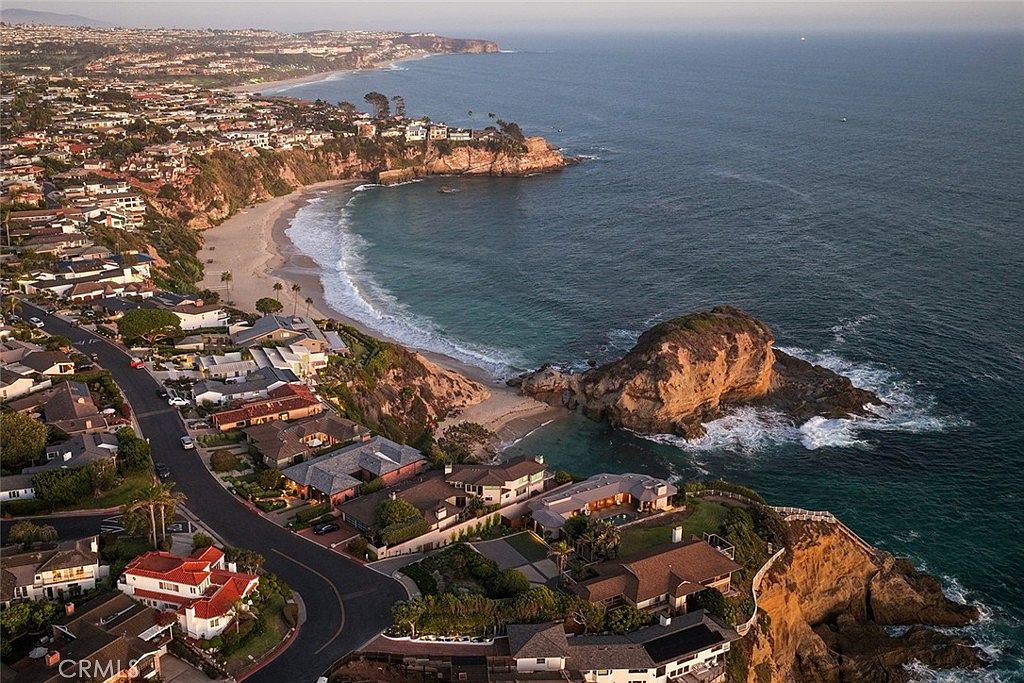 This aerial shot showcases a stunning coastal neighborhood with luxurious homes nestled along the cliffs overlooking the ocean. The turquoise water contrasts beautifully with the sandy beaches and rocky outcrops, creating a picturesque scene. The winding road adds a sense of connectivity to the community.