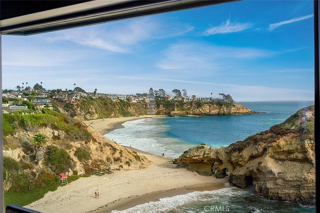 This image showcases a stunning coastal view from a patio or balcony. The scene captures a sandy beach, turquoise ocean waters, and rocky cliffs, creating a serene and luxurious atmosphere. The presence of nearby houses suggests a desirable residential location with premium oceanfront access.