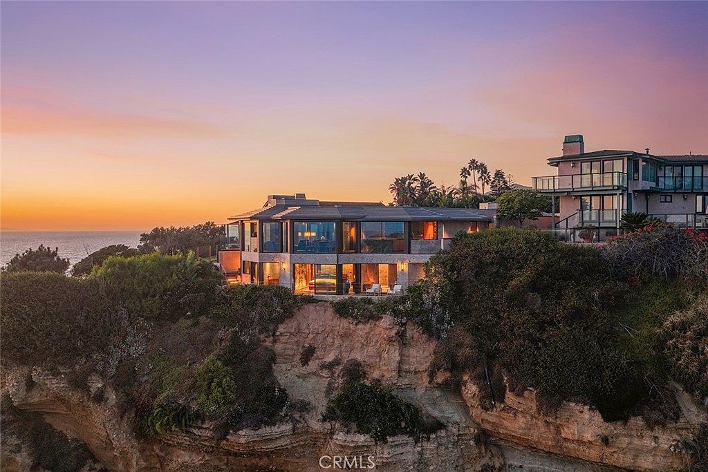 This image showcases a stunning oceanfront property at sunset, featuring modern architecture with expansive glass windows that offer panoramic views. The house is perched on a cliff, surrounded by lush greenery, creating a dramatic and luxurious setting. The warm interior lighting contrasts beautifully with the cool tones of the twilight sky, emphasizing the home's inviting atmosphere.