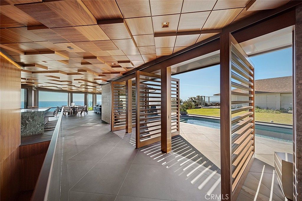 This interior shot showcases a modern hallway with unique architectural details, including a geometrically patterned wooden ceiling and slatted wooden partitions that open to an outdoor pool area. The hallway features large format tile flooring and leads to a dining area with ocean views, creating a seamless transition between indoor and outdoor living spaces. The design emphasizes natural light and a connection to the surrounding environment.