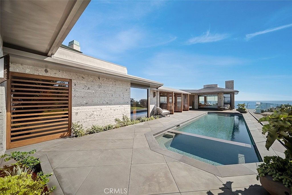 This image showcases a luxurious outdoor pool and spa area of a modern home. The pool features a unique design with a shallow lounging area, surrounded by sleek gray tiling that extends to the house. The architecture is contemporary, with clean lines and a stone facade, complemented by wooden slat accents and lush greenery, creating a serene and upscale ambiance.