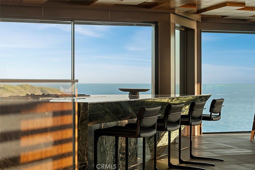 This interior shot showcases a modern kitchen with a stunning ocean view. The kitchen features a marble countertop bar with sleek, black bar stools. Large windows frame the ocean, creating a bright and airy atmosphere, while the wooden ceiling adds a touch of warmth.