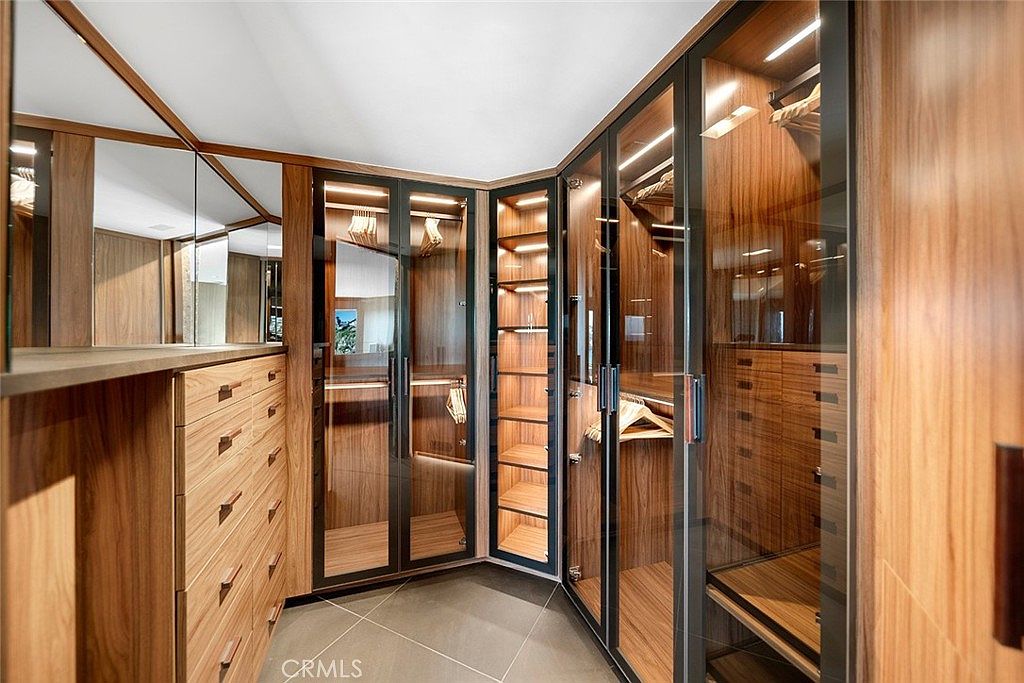 This is a well-organized walk-in closet featuring custom wood cabinetry with glass doors and built-in lighting. The closet includes drawers, shelves, and hanging rods, providing ample storage space. The design is modern and luxurious, enhancing the appeal of the primary suite.