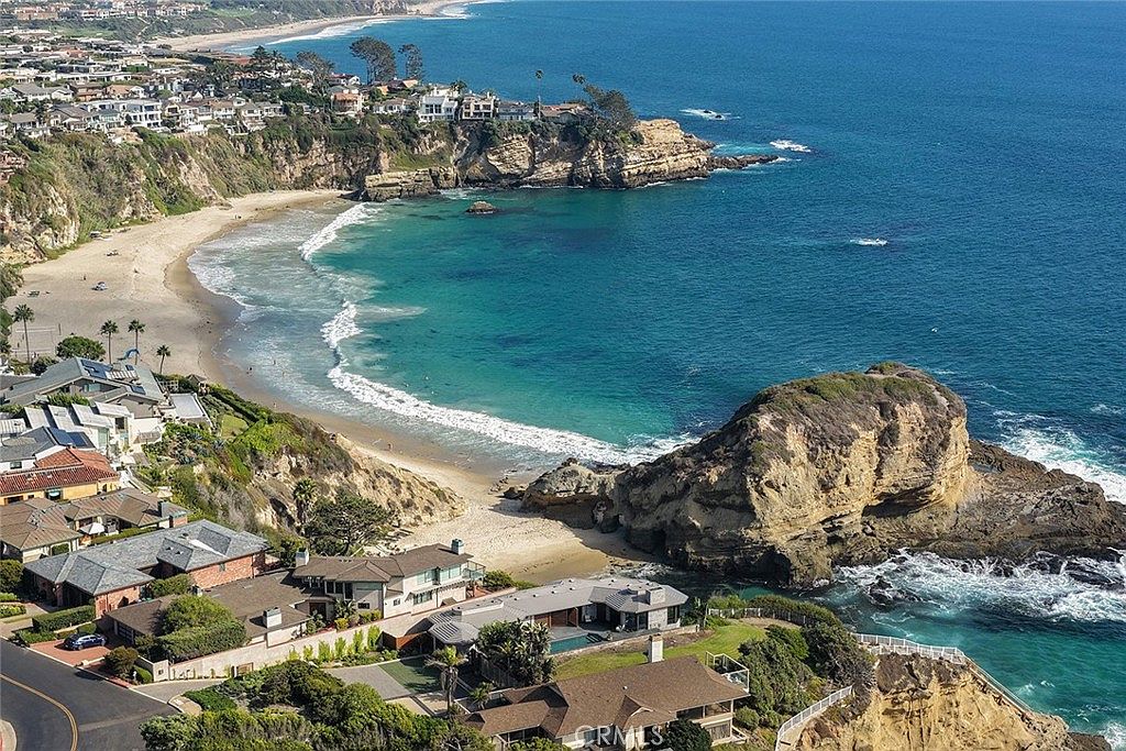 This aerial view showcases a stunning coastal property with a blend of luxury homes nestled along the cliffs overlooking a turquoise ocean. The scene features sandy beaches, rocky outcrops, and lush greenery, creating a picturesque and desirable setting. The overall impression is one of exclusivity and natural beauty, highlighting the prime location and upscale lifestyle.