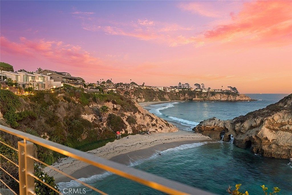 This image showcases a stunning coastal property at sunset, viewed from a patio or balcony. The scene features a sandy beach, turquoise water, and dramatic rock formations, all bathed in the warm hues of the setting sun. Upscale homes are visible on the cliffs above, suggesting a luxurious and desirable location.