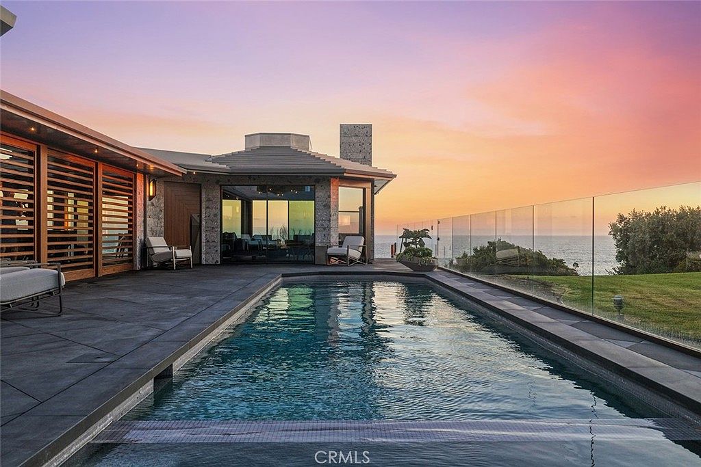 This image showcases a luxurious outdoor pool area at sunset. The pool is surrounded by a modern patio and a glass fence offering unobstructed ocean views. The architecture of the adjacent building features clean lines and natural materials, creating a sophisticated and serene atmosphere.