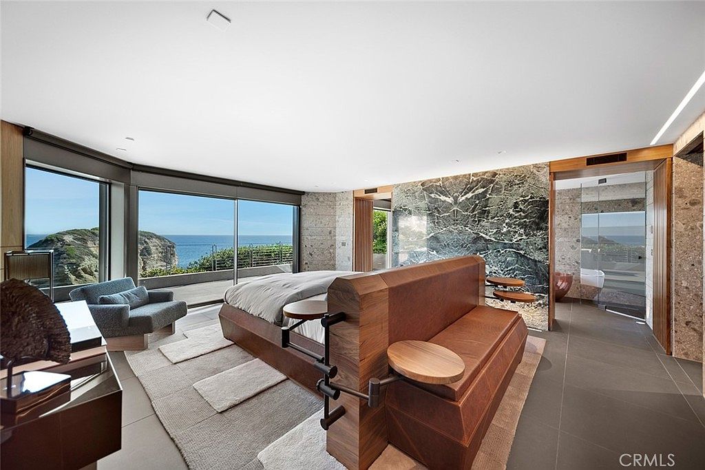 This is a luxurious primary bedroom featuring a unique bed design with integrated side tables and a seating area. The room boasts floor-to-ceiling windows with ocean views, a marble accent wall, and a modern bathroom with glass enclosure. The overall impression is one of high-end comfort and contemporary style.