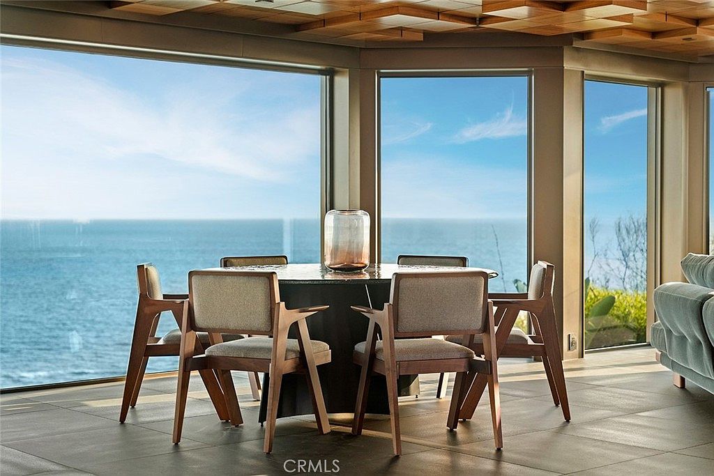 This dining room features a modern aesthetic with a round table surrounded by six chairs with wooden frames and neutral-toned upholstery. Large windows offer a stunning ocean view, enhancing the room's appeal and creating a serene atmosphere. The room is well-lit, highlighting the clean lines and contemporary design.