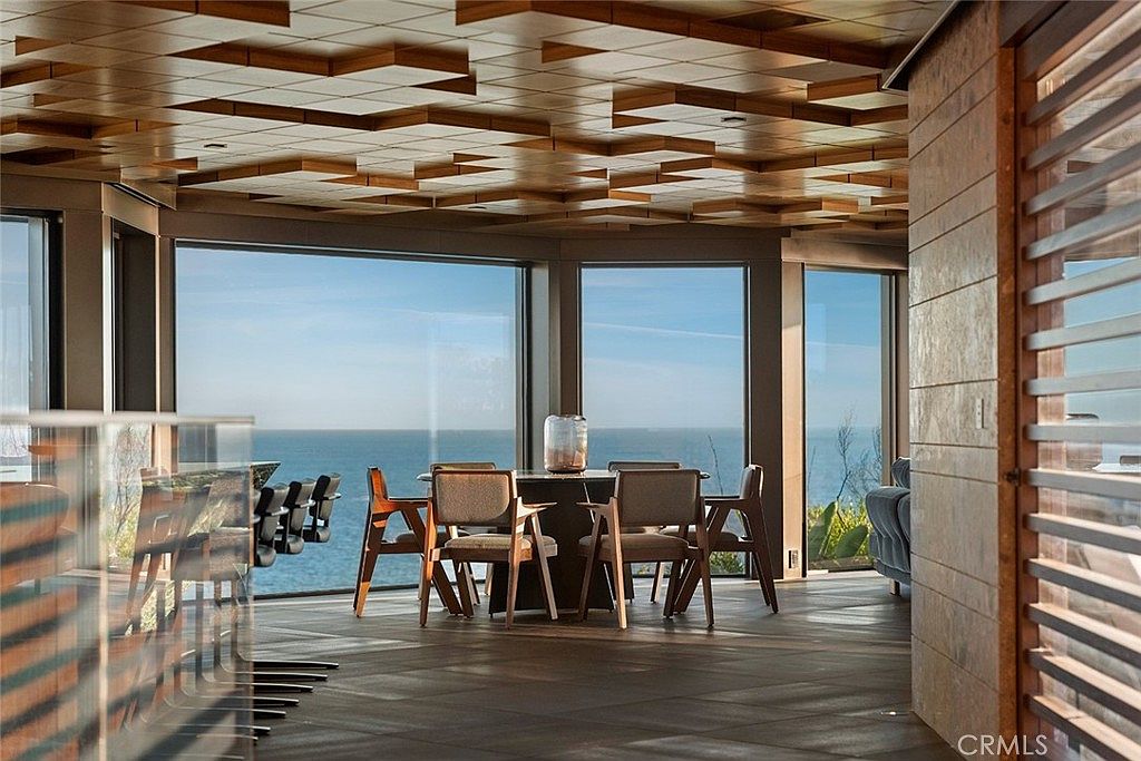 This interior shot showcases a modern dining room with a stunning ocean view through large windows. The room features a unique geometric wood ceiling, a dining table with six chairs, and dark wood flooring. The design emphasizes natural light and a seamless connection to the outdoor scenery, creating an elegant and serene dining experience.