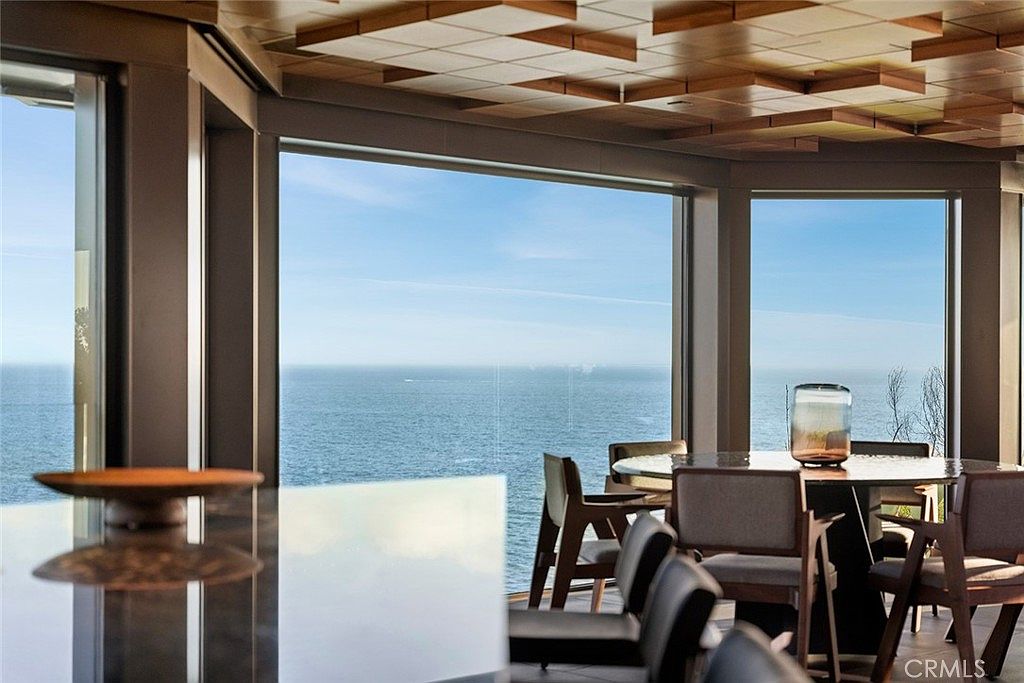 This interior shot showcases a dining area with a stunning ocean view through large windows. The room features a modern aesthetic with a unique wooden ceiling design, a round dining table with chairs, and a decorative glass vase. The overall impression is luxurious and serene, emphasizing the property's prime location and elegant design.