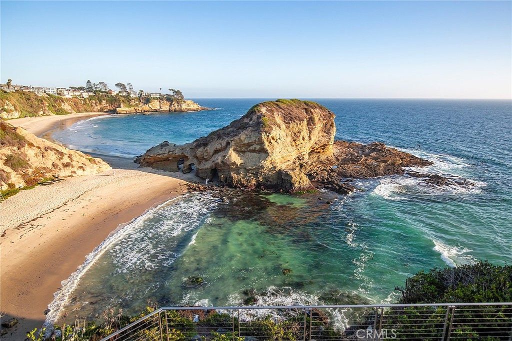 This aerial view showcases a stunning coastal property with a sandy beach, turquoise waters, and a prominent rock formation. The coastline features a mix of sandy beaches and rocky cliffs, with residential buildings visible in the distance. The clear blue sky enhances the overall appeal, creating a picturesque and desirable location.