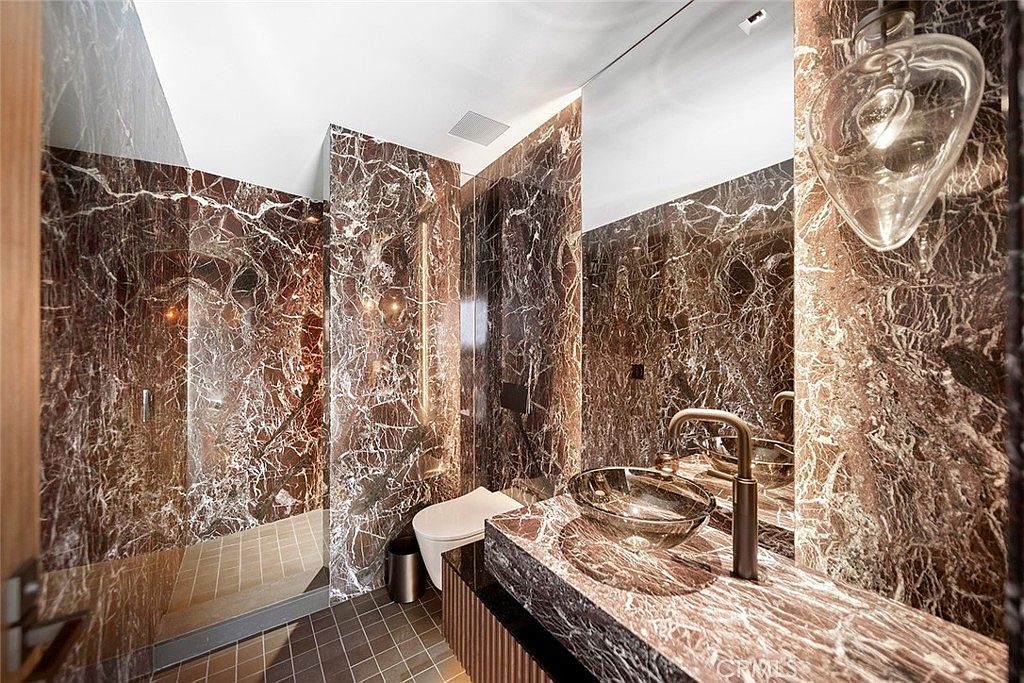 This is a luxurious primary bathroom featuring marble walls and countertops with striking veining. The bathroom includes a modern glass vessel sink, a sleek faucet, and a unique heart-shaped pendant light. A walk-in shower and toilet are also visible, creating a cohesive and high-end aesthetic.