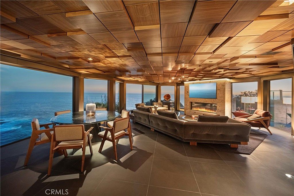 This is a luxurious living room featuring a unique wooden paneled ceiling and expansive windows offering ocean views. The room includes a comfortable sofa, a modern fireplace with a TV above, and a dining area with a glass-top table. The overall impression is one of sophisticated coastal living.