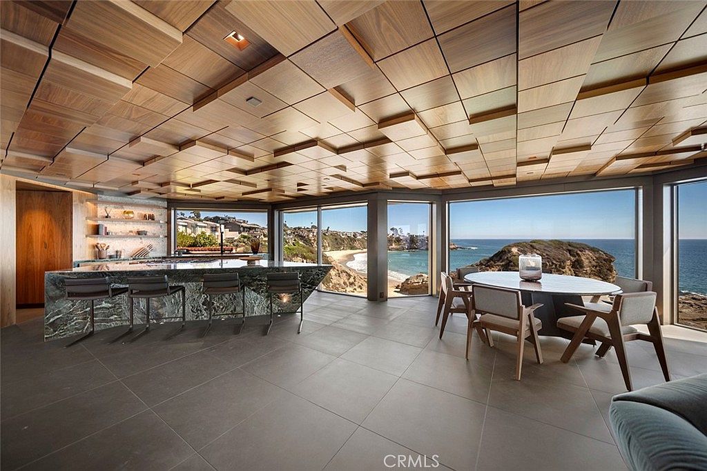 This is a modern kitchen with a unique wooden paneled ceiling and large windows offering ocean views. The kitchen features a green marble countertop island with bar stools and a dining area with a round table and chairs. The overall impression is luxurious and contemporary, emphasizing the stunning coastal location.