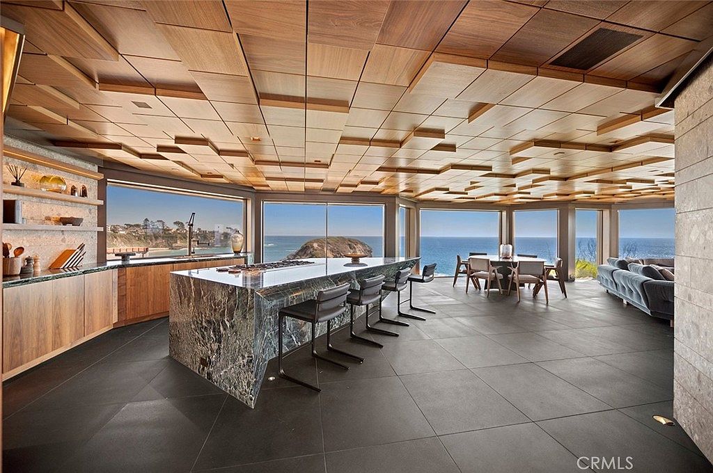 This is a luxurious kitchen and dining area with a stunning ocean view. The kitchen features a large marble island with bar seating, wooden cabinetry, and open shelving. The dining area includes a modern table and chairs, while a comfortable sofa sits nearby, all under a unique wooden paneled ceiling. The space is open and airy, with large windows showcasing the beautiful coastal scenery.