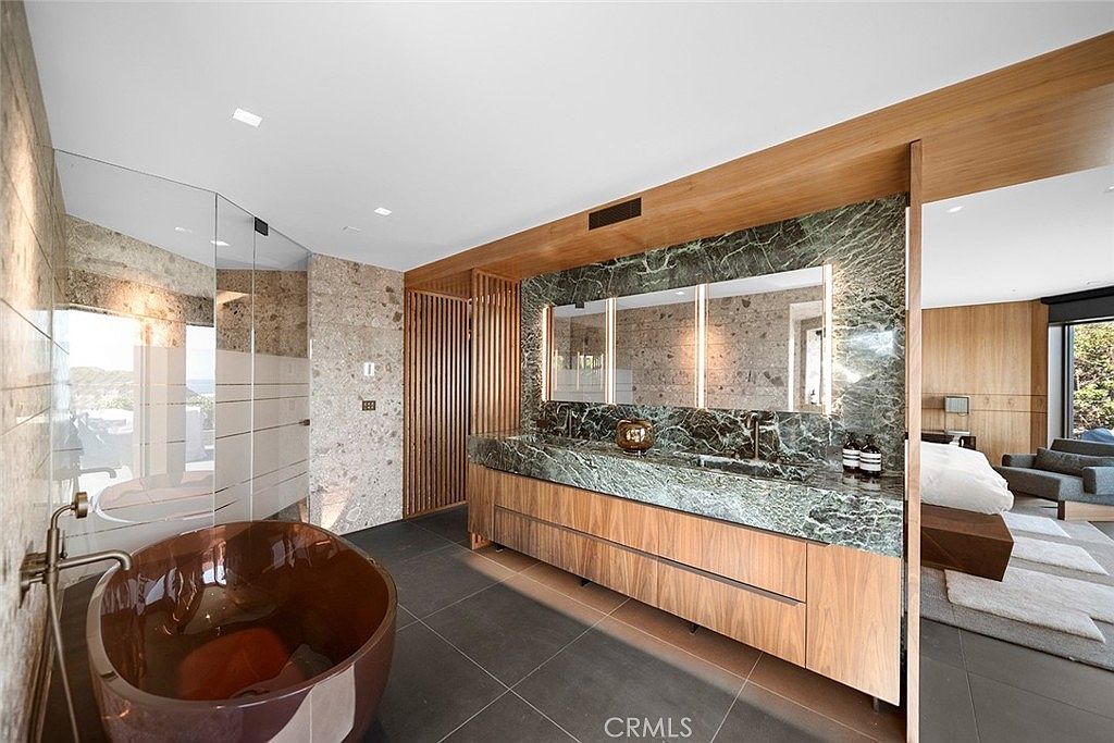 This is a luxurious primary bathroom featuring a modern design with high-end finishes. The bathroom includes a freestanding copper bathtub, a double vanity with a green marble countertop and backsplash, and a separate shower area with glass enclosure. The space is well-lit and opens into a bedroom area, creating a spa-like atmosphere.