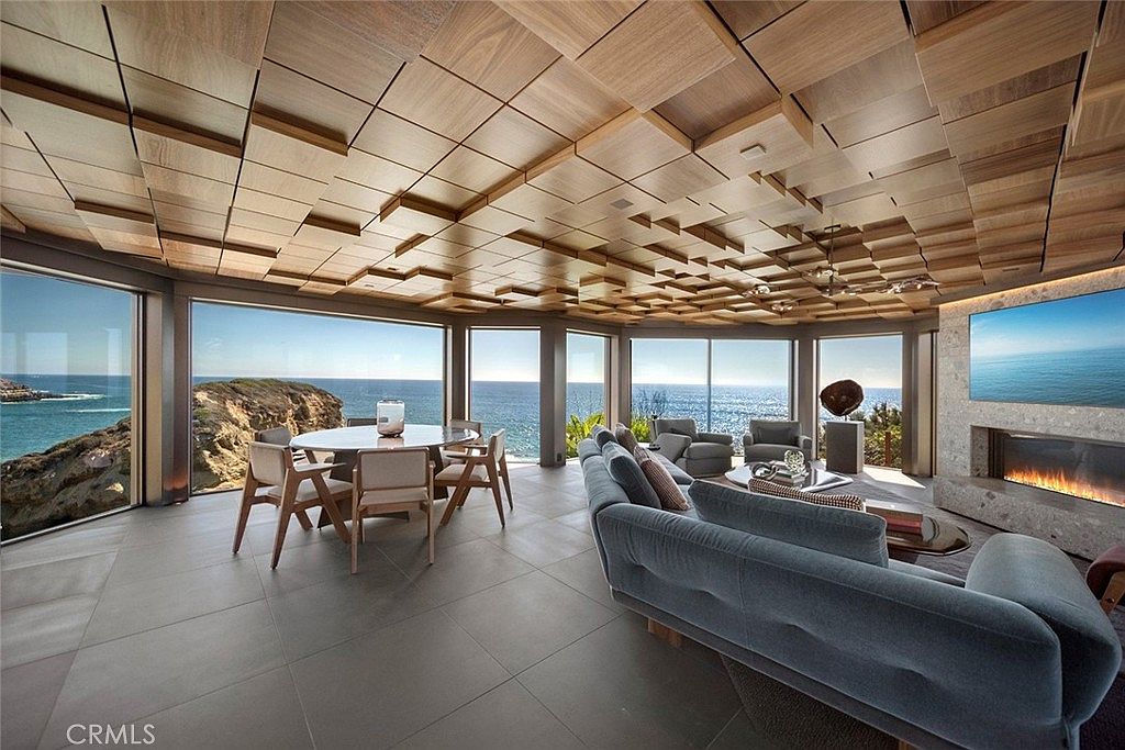 This is a luxurious living room with floor-to-ceiling windows offering stunning ocean views. The room features a unique, geometrically patterned wooden ceiling, a modern fireplace, and stylish, comfortable seating. The open layout seamlessly integrates a dining area, creating a spacious and elegant living space.
