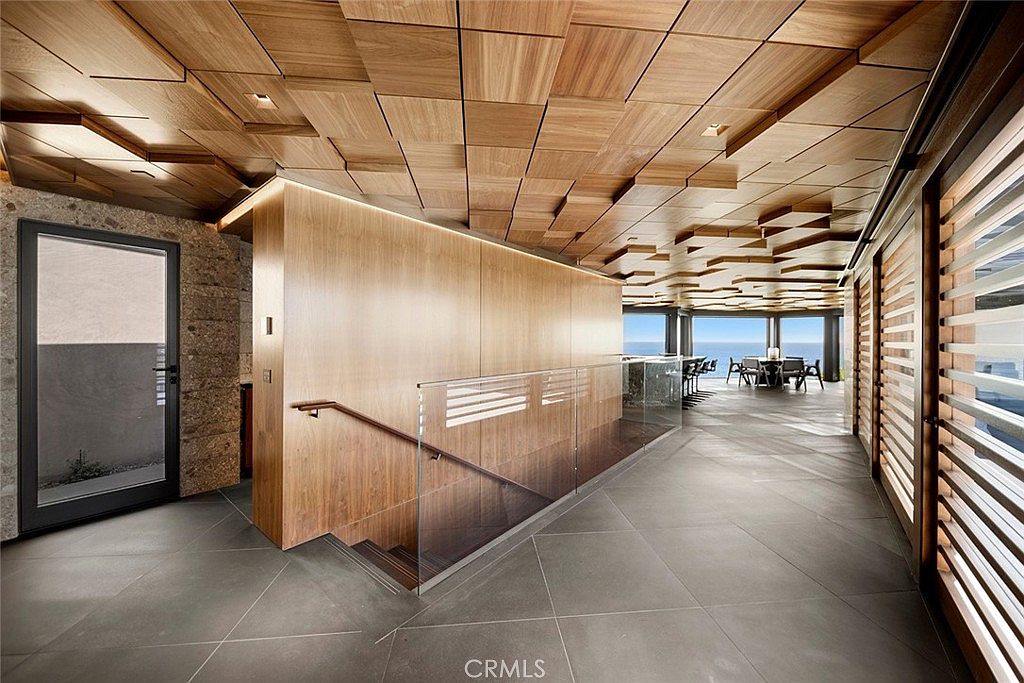 This interior shot showcases a modern hallway with unique architectural details. The ceiling features a geometric wood panel design, complemented by a wooden wall with integrated lighting and a glass railing staircase. Large windows with slatted blinds offer natural light and a glimpse of the ocean view, creating a luxurious and contemporary atmosphere.