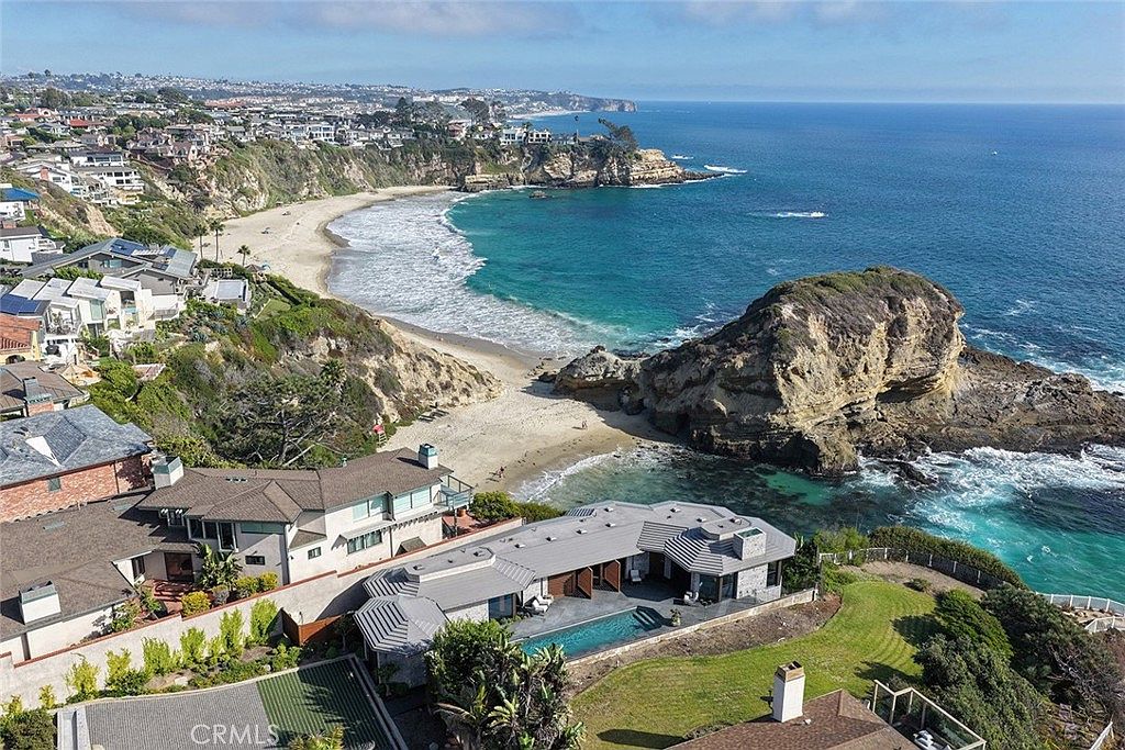 This aerial shot showcases a stunning coastal property with a modern architectural design, featuring a flat roof, a private pool, and meticulously landscaped grounds. The house is perched on a cliff overlooking a picturesque beach and the turquoise ocean, with dramatic rock formations adding to the scenic beauty. The image emphasizes the property's prime location and luxurious outdoor living spaces.