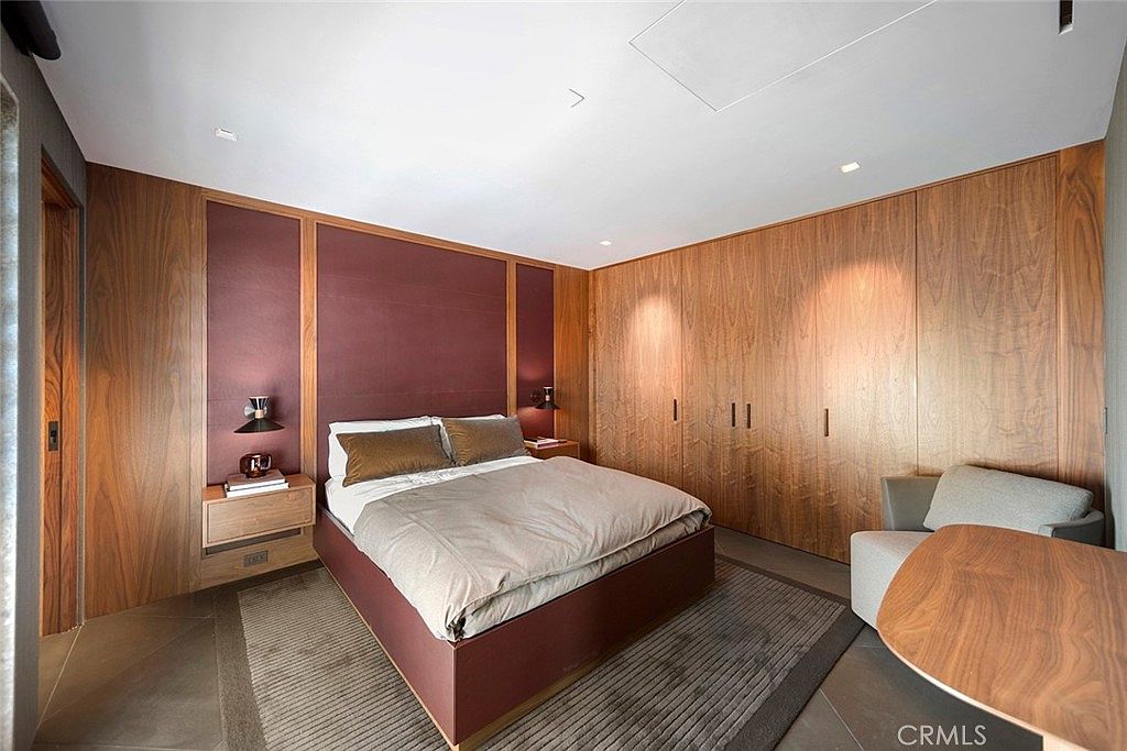 This is a primary bedroom featuring a king-sized bed with a burgundy frame and neutral bedding. The room has wood paneled walls and a built-in wardrobe, creating a warm and luxurious atmosphere. A modern nightstand with a lamp sits beside the bed, and a gray armchair and wooden table are visible in the corner.