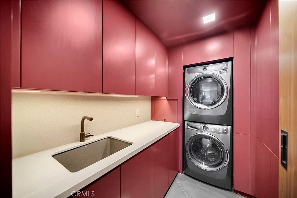 This is a modern laundry room featuring sleek, reddish-pink cabinetry and a stacked washer and dryer unit. A white countertop with a built-in sink and a bronze faucet provides ample workspace. The room is well-lit, creating a clean and efficient space.
