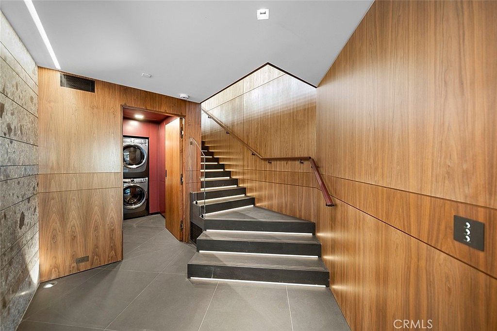 This interior shot showcases a modern hallway with a staircase featuring illuminated steps and a glass railing. The walls are clad in wood paneling, creating a warm and sophisticated ambiance. A laundry room is visible through an open doorway, adding a practical element to the design.