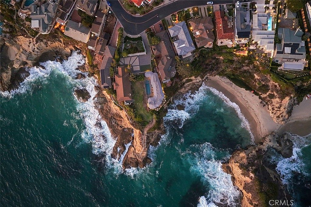 This aerial shot showcases a stunning coastal property with luxury homes perched on cliffs overlooking the ocean. The turquoise water crashes against the rocky shoreline, creating a dramatic contrast with the manicured lawns and swimming pools of the residences. A sandy beach is nestled between the cliffs, offering a private retreat.