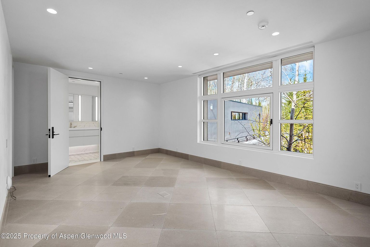 This is a spacious primary bedroom featuring neutral-toned tile flooring and white walls, creating a bright and airy atmosphere. A large window offers natural light and a view of the outdoors, while an open doorway leads to an en-suite bathroom. The room's minimalist design and clean lines contribute to a modern and serene aesthetic.