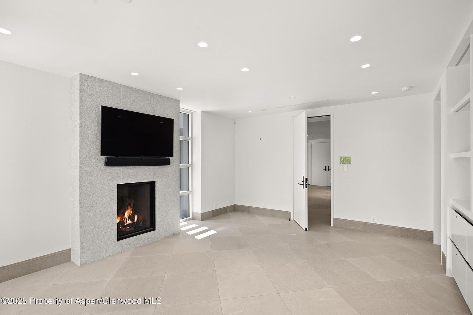 This is an interior shot of a modern living room featuring a fireplace with a large television mounted above it. The room has a neutral color palette with light tile flooring and white walls, creating a bright and airy atmosphere. An open doorway leads to another room, suggesting a spacious and interconnected layout.