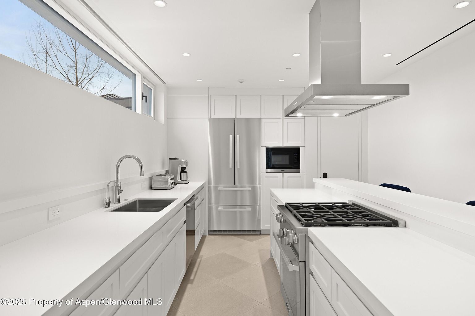 This is a bright, modern kitchen featuring white cabinetry, stainless steel appliances, and light-colored countertops. The kitchen includes a large sink, a stainless steel refrigerator, a microwave, and a gas stovetop with a stainless steel range hood. The room is well-lit with recessed lighting and natural light from a window above the sink, creating a clean and inviting atmosphere.