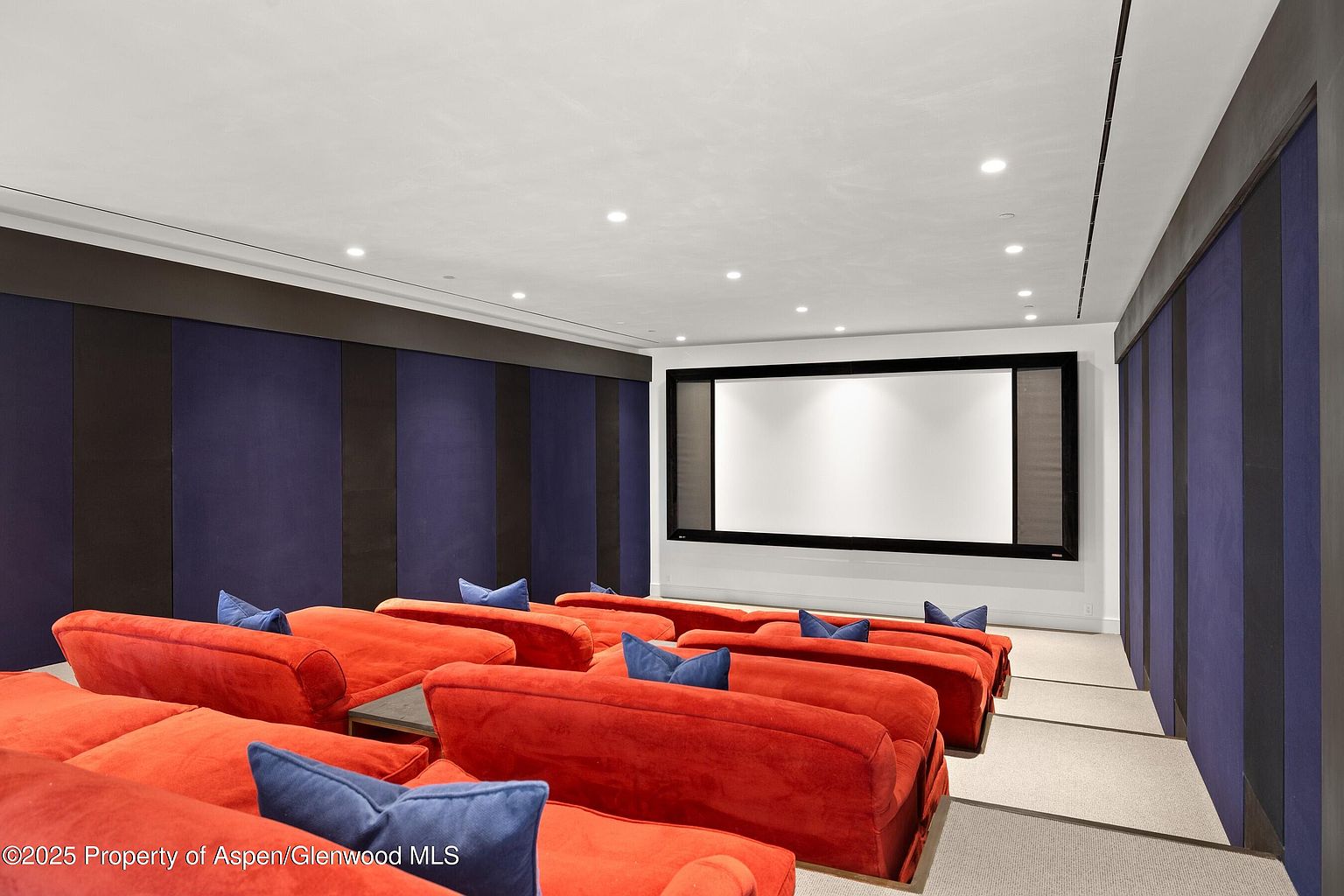 This is an interior shot of a home theater. The room features tiered seating with red couches and blue pillows, a large projection screen, and dark blue and black striped wall panels. The overall impression is one of comfort and luxury, designed for an immersive viewing experience.