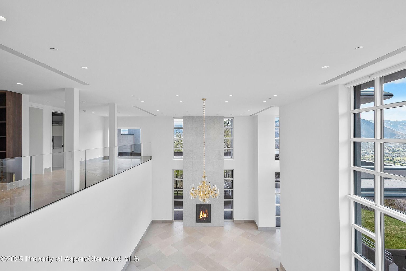 This is a bright and spacious living room featuring a high ceiling and a modern fireplace. The room is illuminated by a large chandelier and natural light streaming through the tall windows, which offer scenic views. The open floor plan and neutral color palette create an airy and inviting atmosphere.