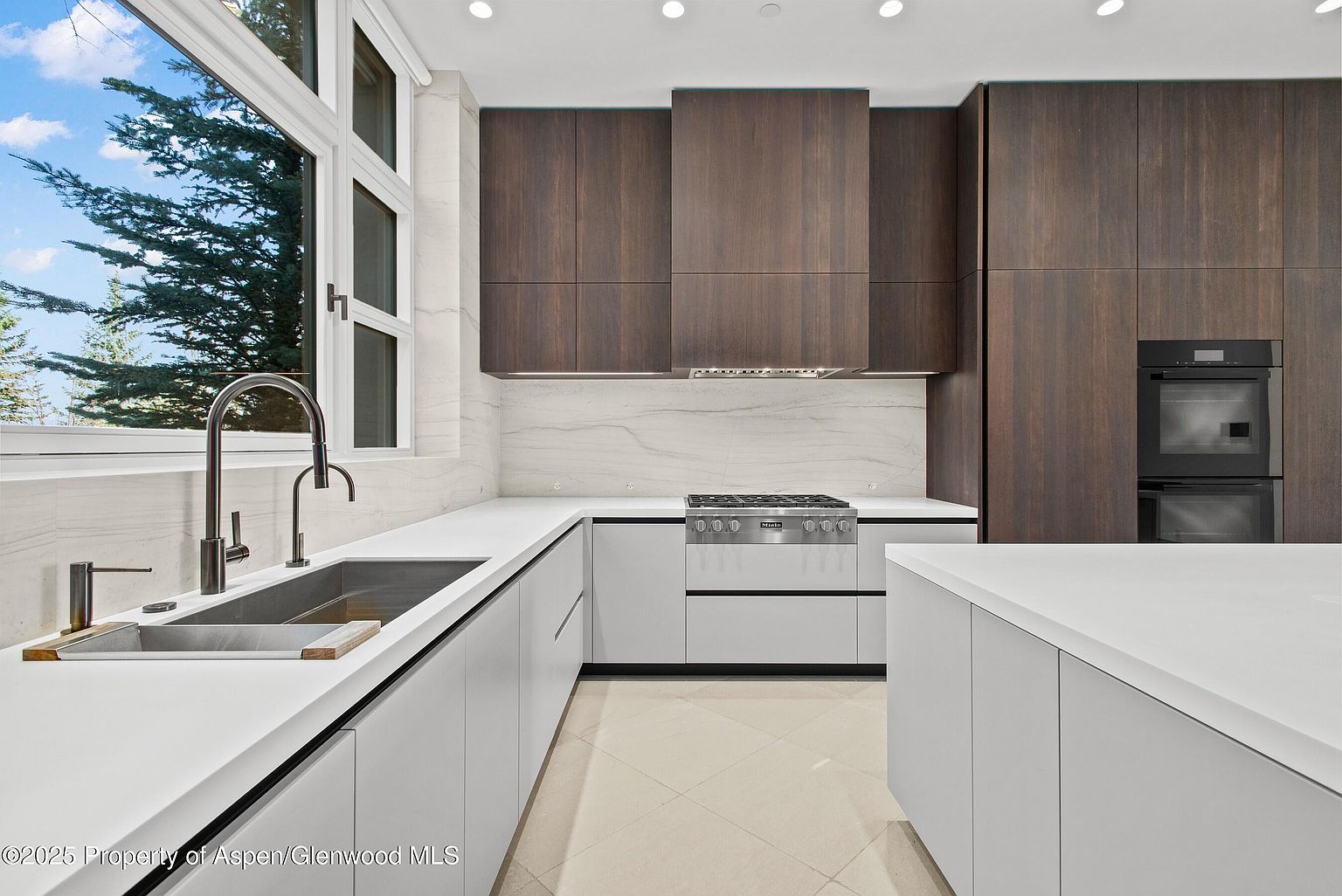 This is a modern kitchen with sleek, handleless white cabinetry and dark wood upper cabinets. The countertops are white, and the backsplash is a light marble. Stainless steel appliances, including a gas range and double oven, are integrated into the design. A large window provides natural light, and an island offers additional counter space. The perspective is from a medium shot, showcasing the entire kitchen layout.