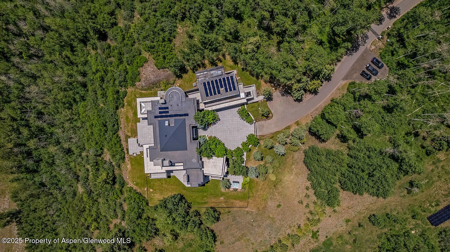 This aerial shot showcases a modern luxury home nestled amidst lush greenery. The property features a flat roof with solar panels, a spacious patio area, and a driveway leading to a parking area with several vehicles. The surrounding landscape is a mix of dense trees and open grassy areas, providing a sense of privacy and seclusion.